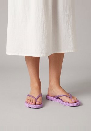 Pies con sandalias de dedo trenzadas moradas y uñas de los pies pintadas a juego, de pie sobre un suelo gris bajo un vestido midi blanco.