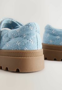 Scarpe in tessuto blu chiaro con design trapuntato, dotate di una suola in gomma marrone testurizzata e delicati motivi floreali bianchi.