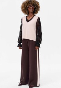 Gebreide mouwloze top in lichtroze, gelaagd over een zwarte doorschijnende blouse met bloemenpatronen. Brede bruine broek met bleek gestreept.