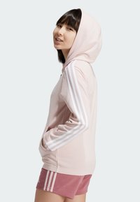 Hoodie rosa claro con capucha ajustable, detalles en blanco con tres franjas en las mangas y bolsillo canguro. Combinado con pantalones cortos deportivos rosas.