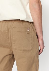 Pantalones de algodón color beige con un corte holgado, que incluyen un bolsillo trasero y una pequeña etiqueta de marca en la costura lateral. Textura suave, diseño minimalista.