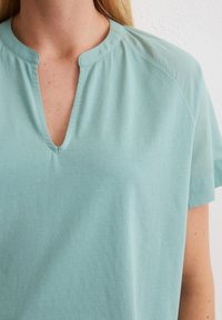 Top verde menta a maniche corte con scollatura a V, tessuto di cotone morbido e dettagli di cucitura discreti lungo la scollatura e le spalle.
