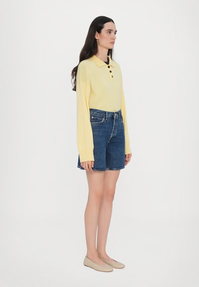 Polo shirt a maglia giallo chiaro con tre bottoni, abbinato a pantaloni corti di jeans blu. Modello indossa scarpe basse beige, in piedi su uno sfondo bianco.