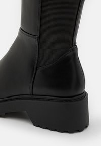 Bottine noire avec un panneau en tissu texturé et en cuir lisse. Elle présente un large talon bloc et une semelle robuste pour une meilleure adhérence.