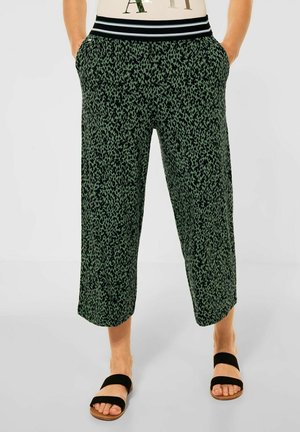 Pantalons coupés à motifs verts avec une taille noire et des poches latérales, présentant un motif léopard en noir contrastant. Chaussures simples.