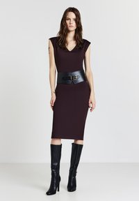 Figurbetontes, dunkelviolettes Kleid mit V-Ausschnitt und kurzen Ärmeln, akzentuiert mit einem breiten schwarzen Ledergürtel mit goldener Schnalle. Schwarze kniehohe Stiefel.