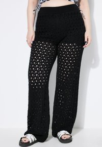 Zwarte gebreide broek met een opengewerkt ontwerp, voorzien van een brede elastische tailleband en een losse pasvorm. Gecombineerd met zwart-witte sandalen.
