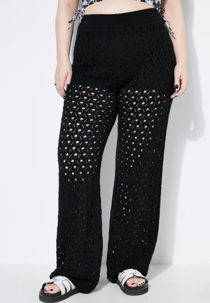 Pantaloni neri a maglia con un design traforato, dotati di una cintura elasticizzata ampia e di una vestibilità morbida. Abbinati a sandali neri e bianchi.