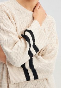 Maglione lavorato a maglia color crema con motivi testurizzati, caratterizzato da strisce orizzontali nere sui polsini. Vestibilità rilassata e scollatura rotonda.