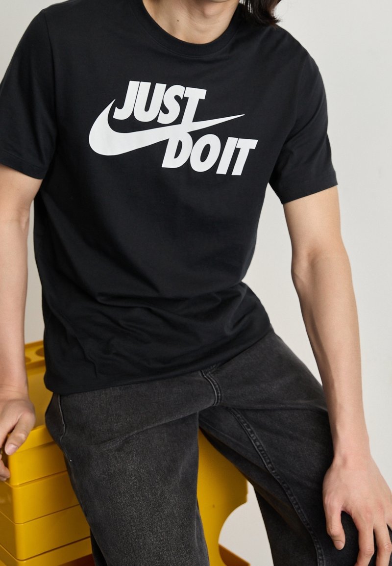Person wearing a black "Just Do It" Nike T-shirt and dark jeans, sitting on a yellow crate against a plain background.