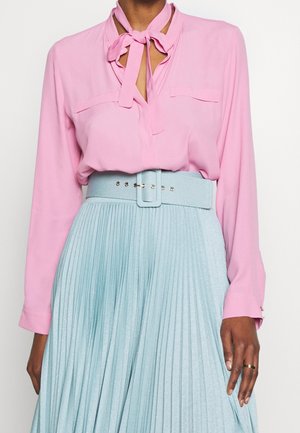 Femme portant un chemisier long à manches roses avec une cravate et une jupe plissée taille haute bleu clair avec une large ceinture.