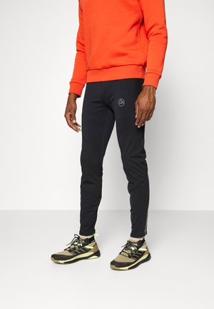 Personne portant un pantalon de sport noir avec un petit logo sur la cuisse, un sweat-shirt orange et des chaussures de sport beige et noir, debout devant un fond blanc.
