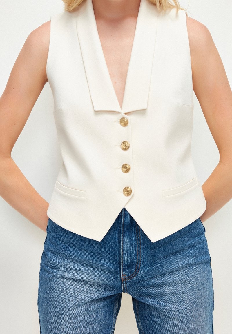 Femme portant un gilet blanc sans manches avec des boutons dorés sur un jean bleu, montrée des épaules aux hanches sur un fond uni.