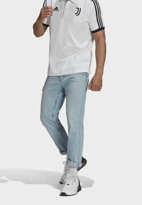 Homme portant un polo blanc Adidas Juventus, un jean bleu clair retroussé, des baskets blanches et des chaussettes blanches, debout sur un fond gris.