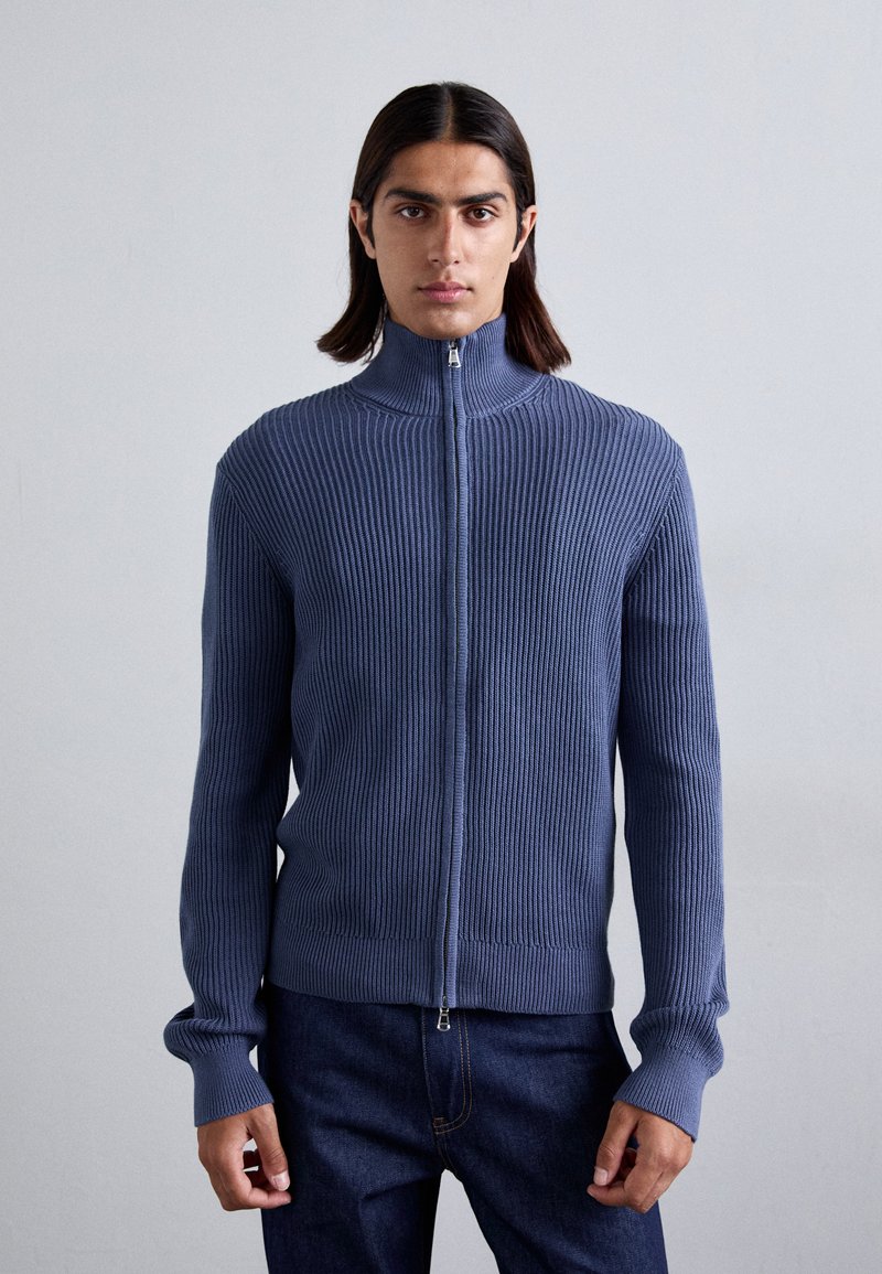 Ριγέ μπλε πουλόβερ με ψηλό φερμουάρ, εφαρμοστή σχεδίαση �και μακριά μανίκια. Διαθέτει λεία υφή και διακριτές λεπτομέρειες ραψίματος.