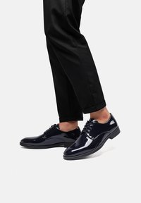 Scarpe eleganti da uomo in pelle verniciata blu navy con finitura lucida, design con lacci, dettagli cuciti e tacco basso. I pantaloni sono neri e su misura.