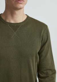 Olivno zeleni pulover s okroglim izrezom, rebrastim ovratnikom in subtilnimi križnimi šivi na ramenskih šivih, izdelan iz mehke, teksturirane tkanine.