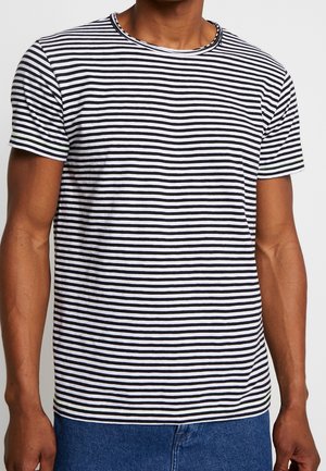 Homme portant un T-shirt à manches courtes rayé horizontalement noir et blanc et un jean bleu, debout les bras détendus.