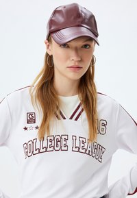 Bordeaux rotes Kunstleder-Cap mit glatter Oberfläche und genähten Details, getragen zu einem weißen Trikot mit dem Text "COLLEGE LEAGUE" in Burgunderrot.