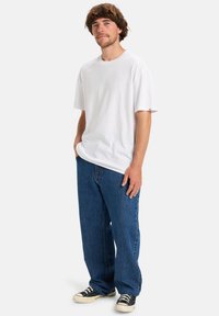 Man met mediumbruin haar die een eenvoudig wit T-shirt, losse blauwe jeans en zwart-witte sneakers draagt, staan met handen in de zakken.