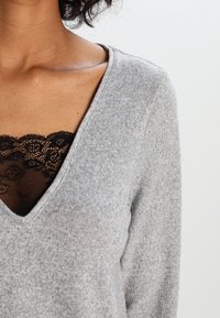 Grauer V-Ausschnitt-Pullover aus weichem Material, mit einem strukturierten Muster, kombiniert mit einem schwarzen Spitzen-Bralette darunter, das teilweise sichtbar ist.