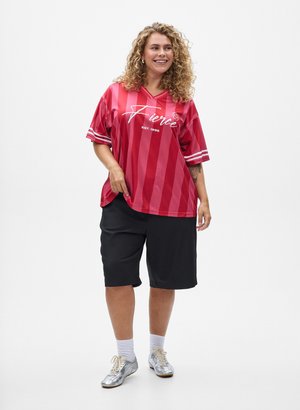 Rødstribet oversized trøje med hvid skrift, parret med sorte shorts. Modellen bærer sølvfarvede sko og hvide sokker.