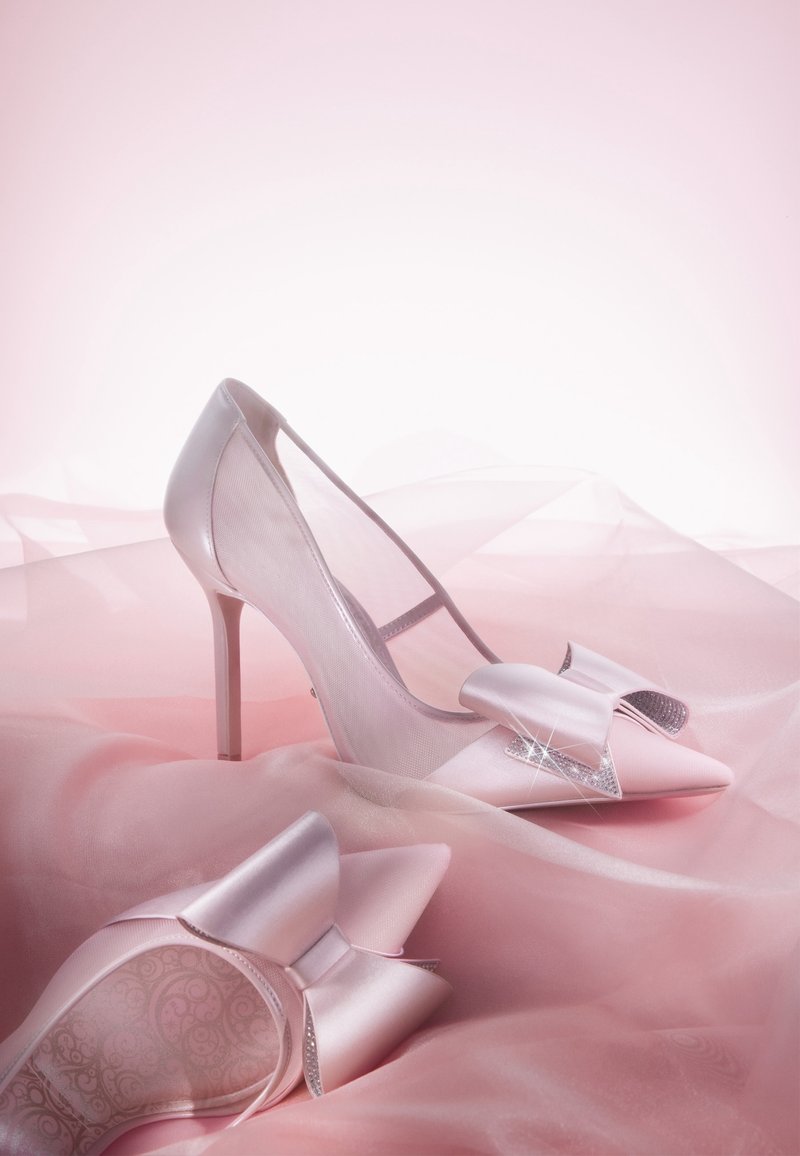 Des chaussures à talons hauts pink avec une finition satinée, un bout pointu, des côtés translucides, et un grand nœud avec des accents en strass à l'avant.