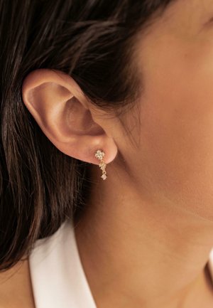 Goldene Ohrringe mit einer floralen Stecker und einer hängenden Kette mit kleinen klaren Edelsteinen, die in der Nähe des Ohrs einer Person präsentiert werden.