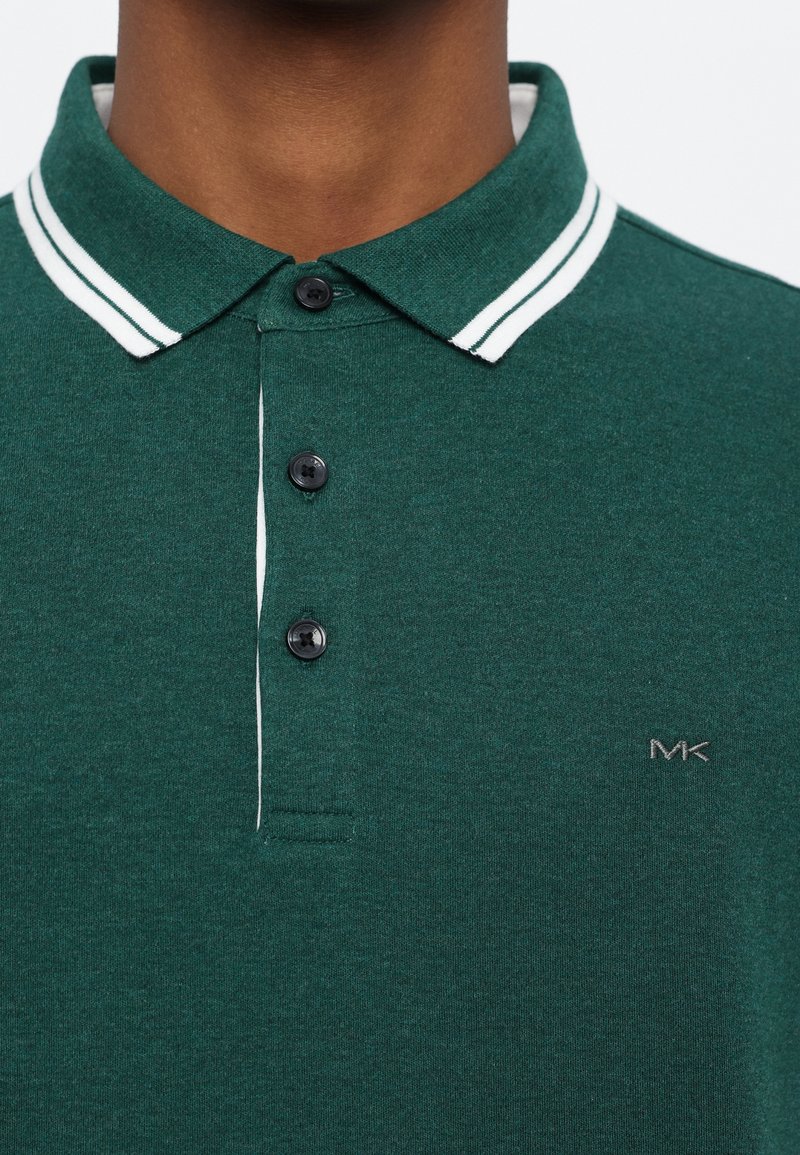 Nahaufnahme eines grünen Poloshirts mit weißen Doppelstreifen am Kragen und drei schwarzen Knöpfen, auf der Brust ist ein kleines "MK"-Logo zu sehen.