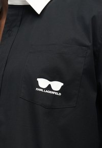 Τσέπη μαύρου πουκάμισου με λευκό γραφικό γυαλιών ηλίου και το κείμενο "KARL LAGERFELD" κάτω από το γραφικό.