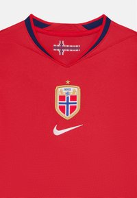 Punane spordisärk tekstureeritud kangast, V-kaelusega, millel on kullast ja sinisest Norra vapp ja valge Nike logo ees.