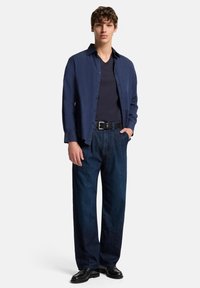 Camicia blu navy a maniche lunghe sopra una t-shirt scura con scollo a V, abbinata a pantaloni a gamba larga in denim scuro e scarpe nere, tutto su sfondo bianco.