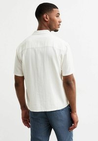 Korte mouwen, licht beige shirt met een kraag. De stof lijkt getextureerd met een ontspannen pasvorm. Draagt met blauwe gestreepte pantalons, zichtbaar vanaf de achterkant.