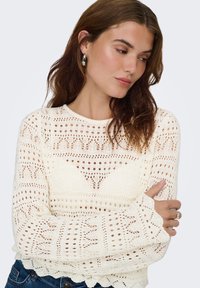Pull en crochet crème avec motifs ouverts, encolure ronde et manches longues, présentant un ourlet festonné et des détails texturés.