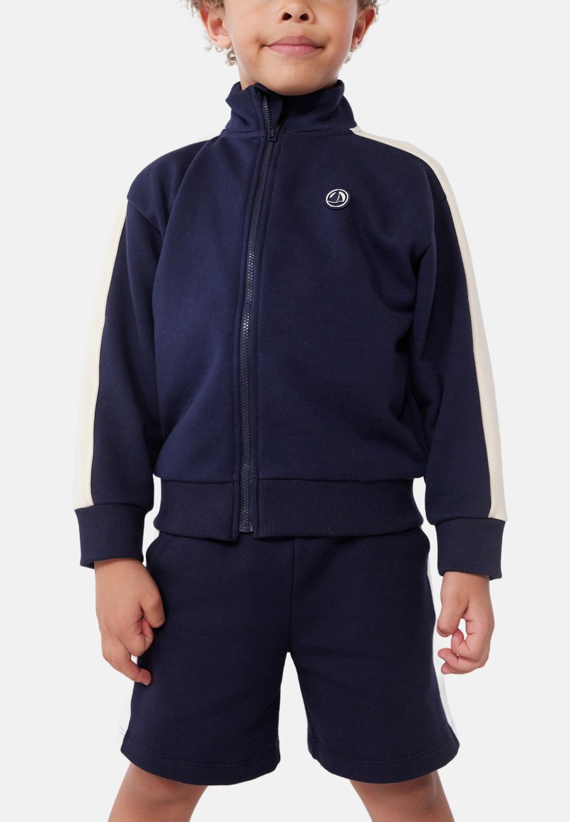 Marineblauer Trainingsanzug mit weißen Seitenstreifen; Reißverschlussjacke mit hohem Kragen, gerippten Bündchen und passender Shorts, aus weichem Stoff gefertigt.
