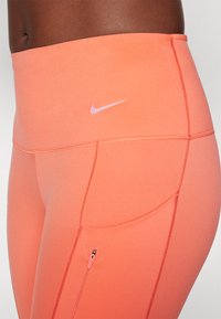Nike leggings i lys korall med en glatt tekstur. Har en glidelåslomme og en liten hvit logo på midjebåndet.