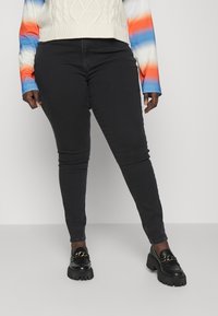 Calças skinny pretas com uma textura suave, combinadas com sapatos loafer pretos robustos com um detalhe em corrente dourada. O conjunto inclui um suéter em blocos de cores.