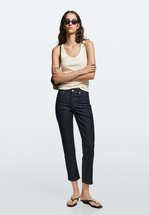 Kvinde iført sorte solbriller, kremfarvet ærmeløs top, mørke korte jeans, sorte klipklapper og en lille skuldertaske, står med armene over kors.