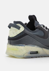 Baskets noires avec une superposition brillante, empeigne en mesh, unité Air visible, et semelle en mousse mouchetée. Détails précis en vert clair et gris.