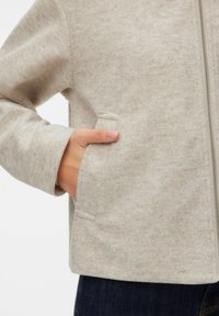 Main dans la poche d'une veste en laine beige à fermeture éclair portée sur un jean bleu foncé.