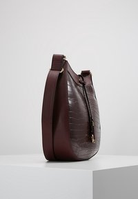 Viininpunainen olkalaukku kiiltävällä krokotiilinkuviolla, kaartuvalla muodolla ja kullanvärisillä metalliosilla; siinä on sivussa hapsukoriste ja tukeva hihna.