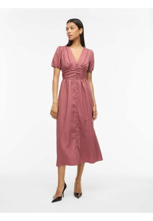 Kvinde iført en mauve midi kjole med pufærmer, knapdetaljer og en dyb V-hals, stående i sorte spidse høje hæle.