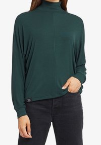 Maglia a maniche lunghe di colore verde scuro con collo alto, vestibilità comoda, texture a costine e orlo che scende sotto la vita. Etichetta visibile in basso.