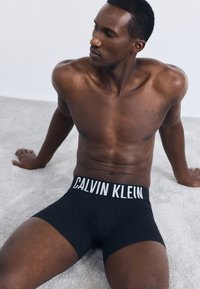 Fekete Calvin Klein boxer alsónadrág logós derékrésszel. A modell izmos testalkatú, egy puha, világos színű felületen ül.