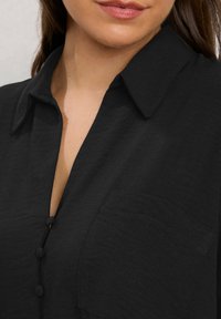 Camicia nera con colletto, chiusura con bottoni e taschino sul petto. Il tessuto è texture con una leggera lucentezza, che mette in risalto la sua superficie liscia.