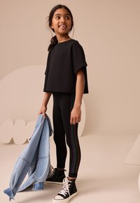 Sort croppet top og sorte leggings med regnbuefarvede nitteaccents, kombineret med en lyseblå denimjakke. Modellen har sorte højtstående sneakers på.
