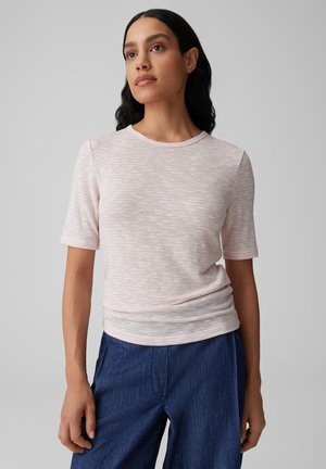 Frau mit langen dunklen Haaren, die ein hellrosa kurzärmliges Oberteil und blaue Hosen trägt, vor einem schlichten grauen Hintergrund stehend.