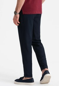 Pantaloni navy a gamba affusolata, con una texture liscia e orli arrotolati, abbinati a scarpe scure e una camicia bordeaux, con dettaglio dell'orologio al polso.