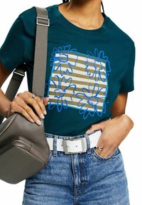 Teal bomullströja med blå blommegrafik och beiga ränder, kombinerad med högmidjade denimshorts och en grå axelväska.