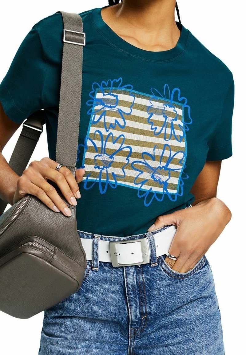 Teal bomullströja med blå blommegrafik och beiga ränder, kombinerad med högmidjade denimshorts och en grå axelväska.